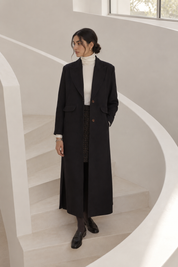 A woman stands on a white spiral staircase wearing a long burgundy coat over a white turtleneck sweater, a dark patterned skirt, and black loafers.