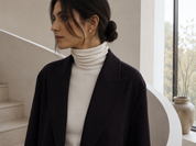 A woman wearing a burgundy tailored coat over a cream ribbed turtleneck sweater, standing indoors near a curved white wall and window.