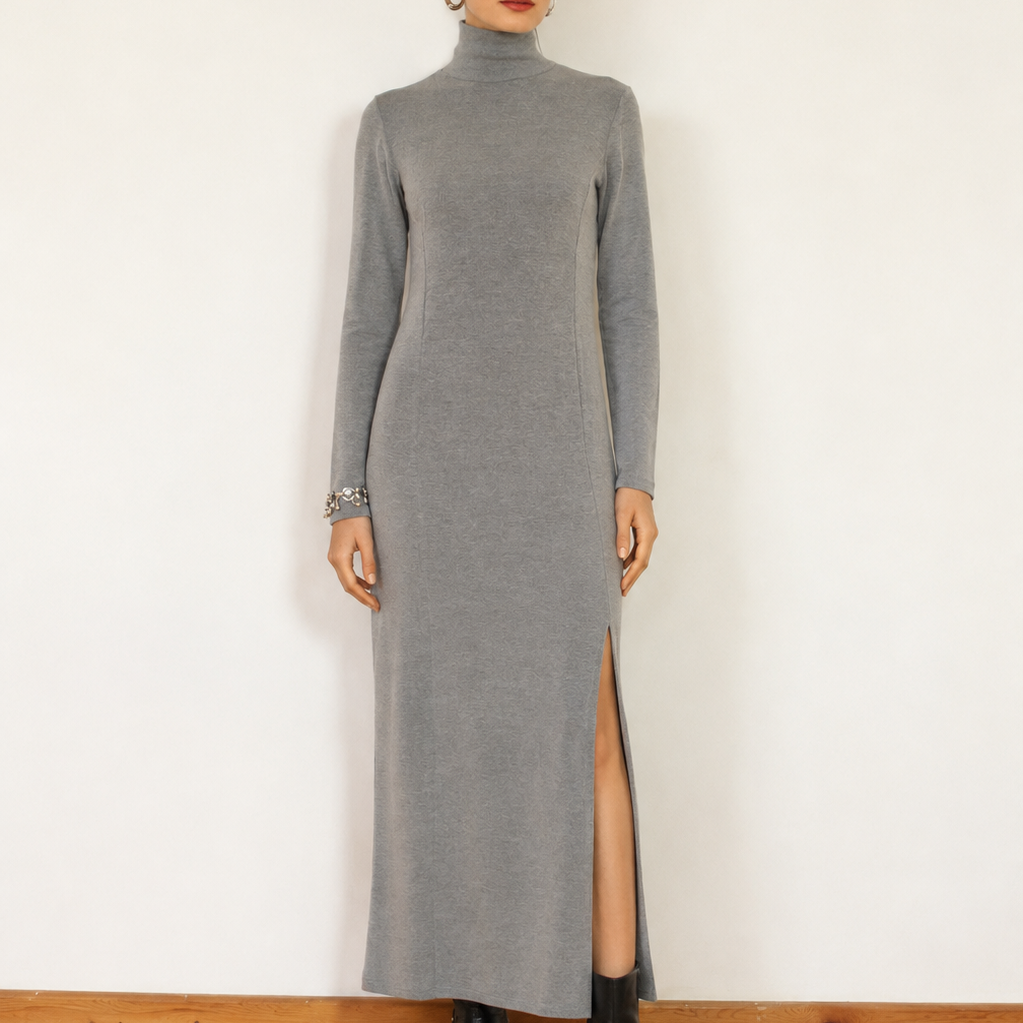 A woman stands against a white wall wearing a long-sleeve gray mock-neck maxi dress with a thigh-high slit, paired with black ankle boots.
