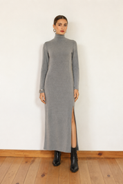 A woman stands against a white wall wearing a long-sleeve gray mock-neck maxi dress with a thigh-high slit, paired with black ankle boots.