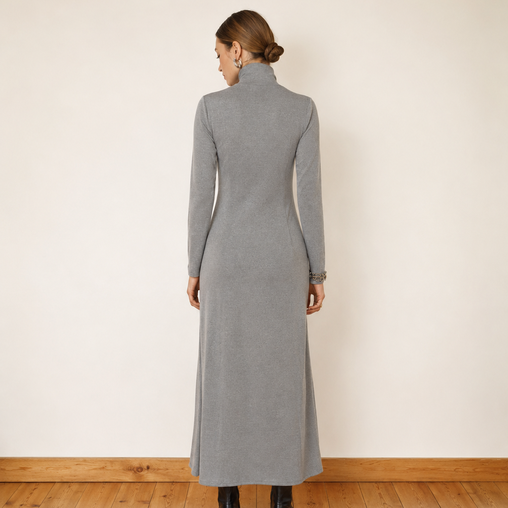 A woman stands with her back to the camera wearing a long-sleeved gray mock-neck midi dress, paired with black ankle boots, in a minimalist room with a cream wall and wooden floor.