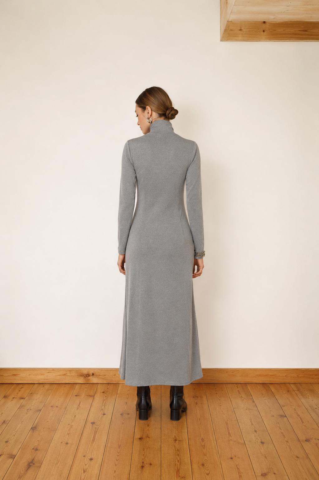 A woman stands with her back to the camera wearing a long-sleeved gray mock-neck midi dress, paired with black ankle boots, in a minimalist room with a cream wall and wooden floor.