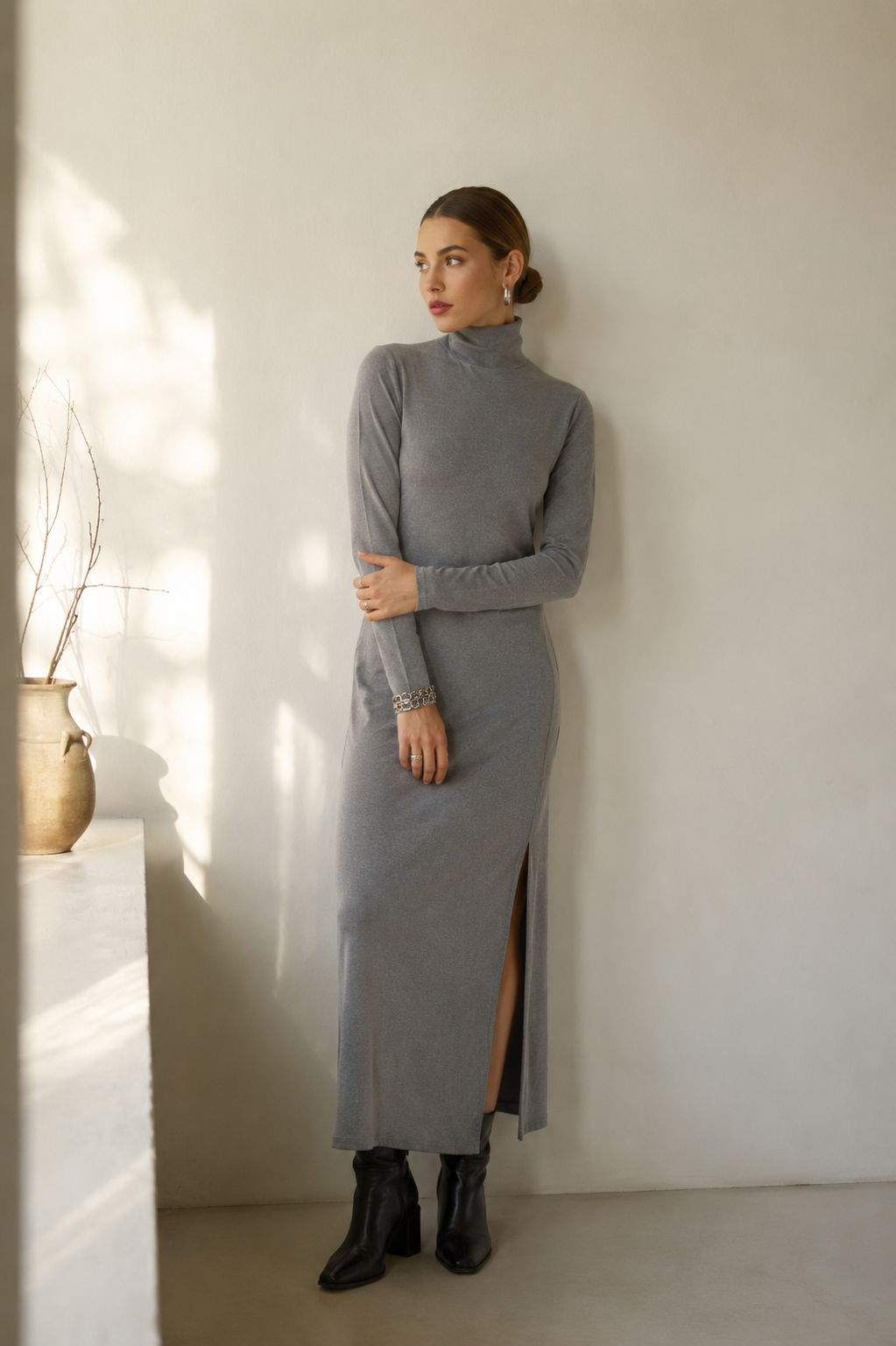 A woman stands in a minimalist room wearing a long-sleeve gray mock-neck midi dress with a side slit, paired with black ankle boots.