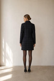 A woman stands with her back to the camera wearing a dark plaid blazer dress, black tights, and black heels in a minimalist studio setting with warm sunlight casting shadows on the wall.