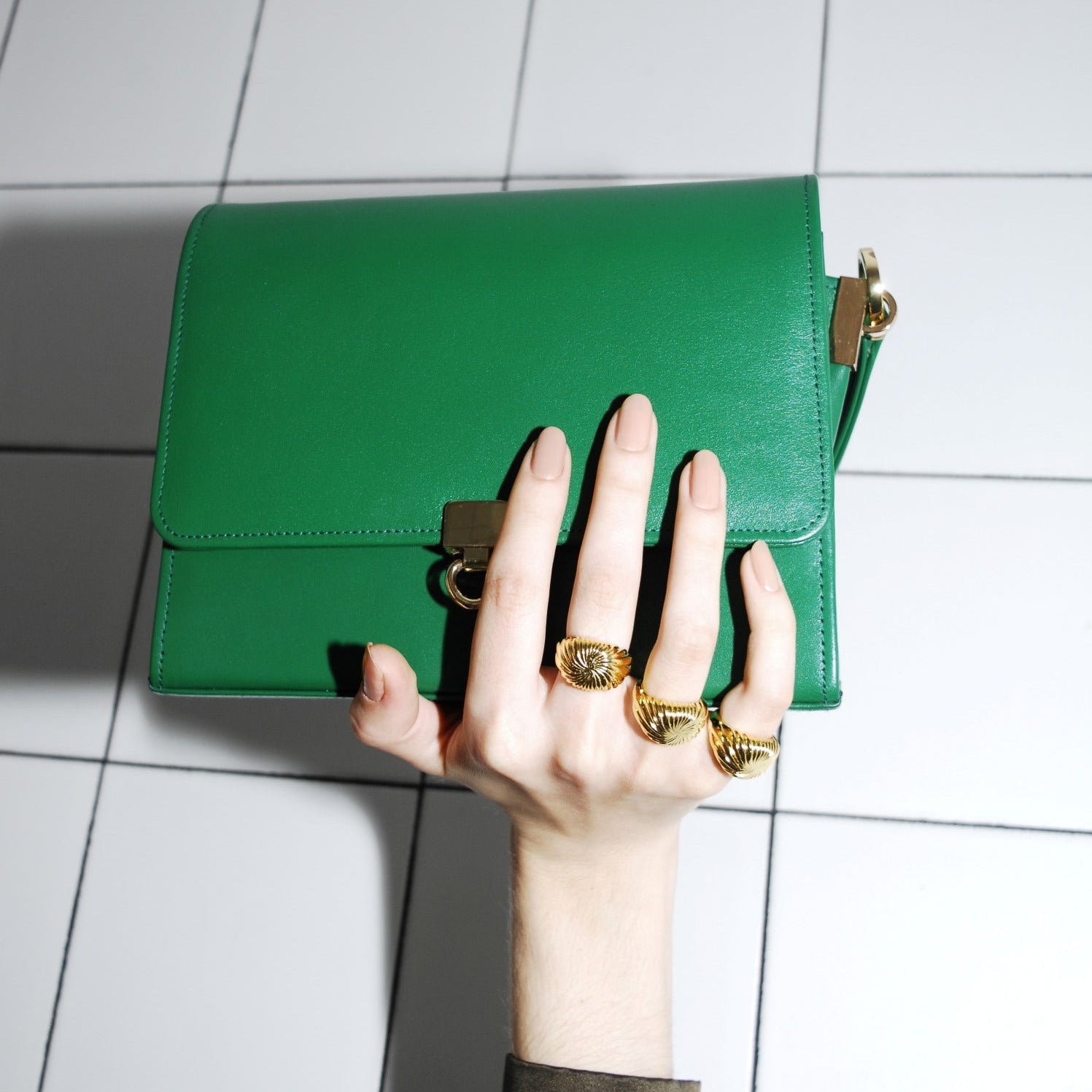 Green handbag held by a hand with gold rings on a tiled floor