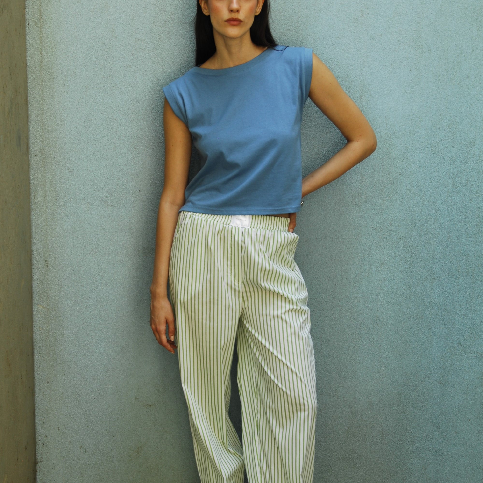 A woman standing wearing green Kyle striped cotton pants with an elasticated waist and side pockets, paired with a Dahlia blue I-shirt.