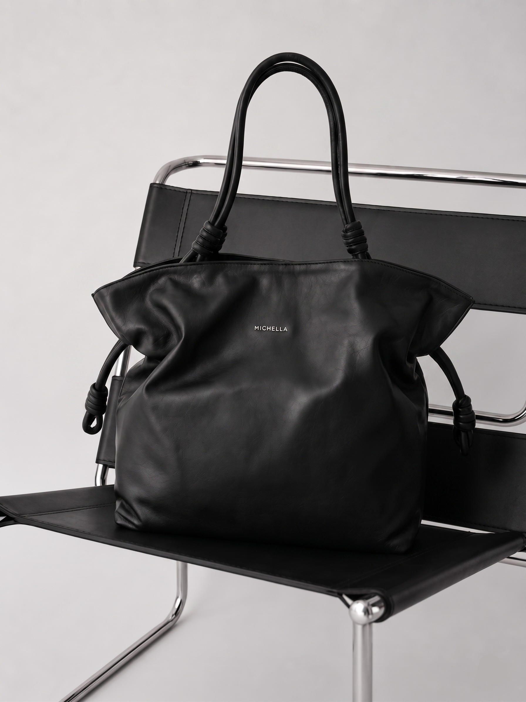 Black handbag with 'Michella' branding on a black chair against a light gray background