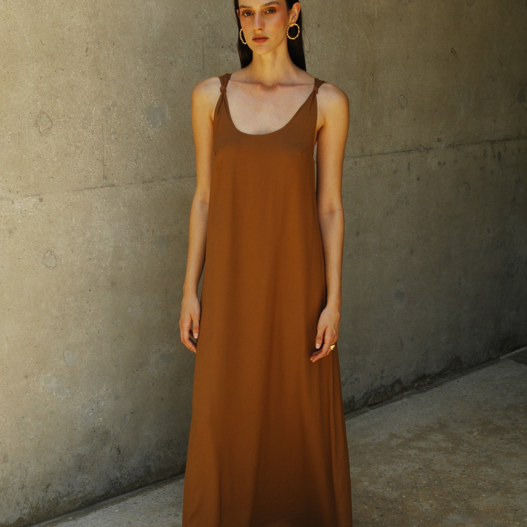 A model wearing a brown maxi dress with a deep neckline and knot straps, standing against a concrete background.
