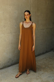A model wearing a brown maxi dress with a deep neckline and knot straps, standing against a concrete background.