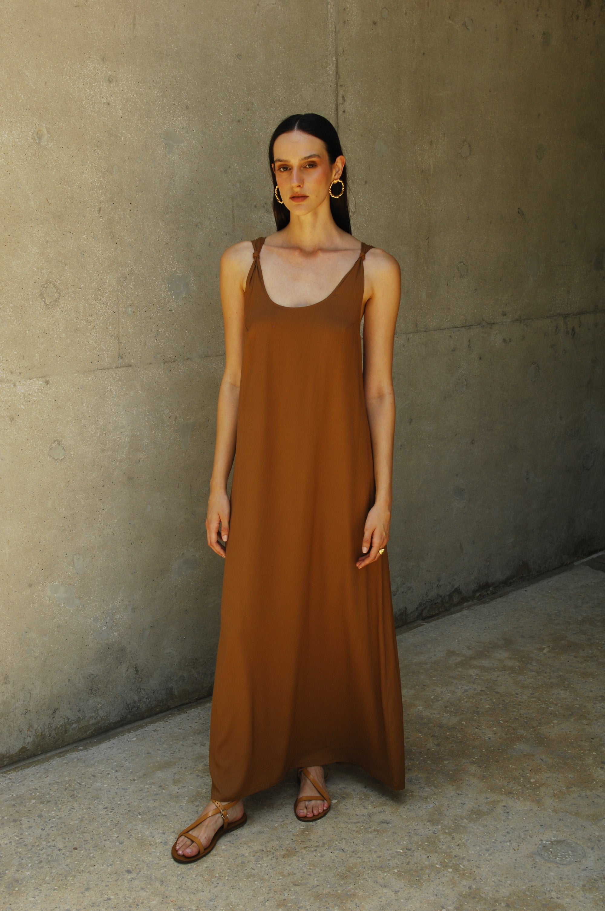 A model wearing a brown maxi dress with a deep neckline and knot straps, standing against a concrete background.