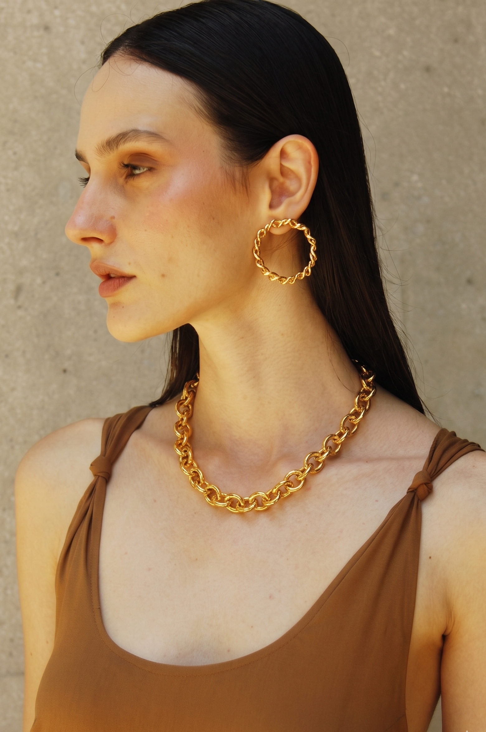 Woman wearing Sia gold hoop earrings and chain necklace against a beige wall