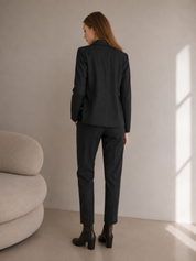 Woman wearing a dark grey suit standing in a minimalistic room with a beige sofa and light-colored wall.