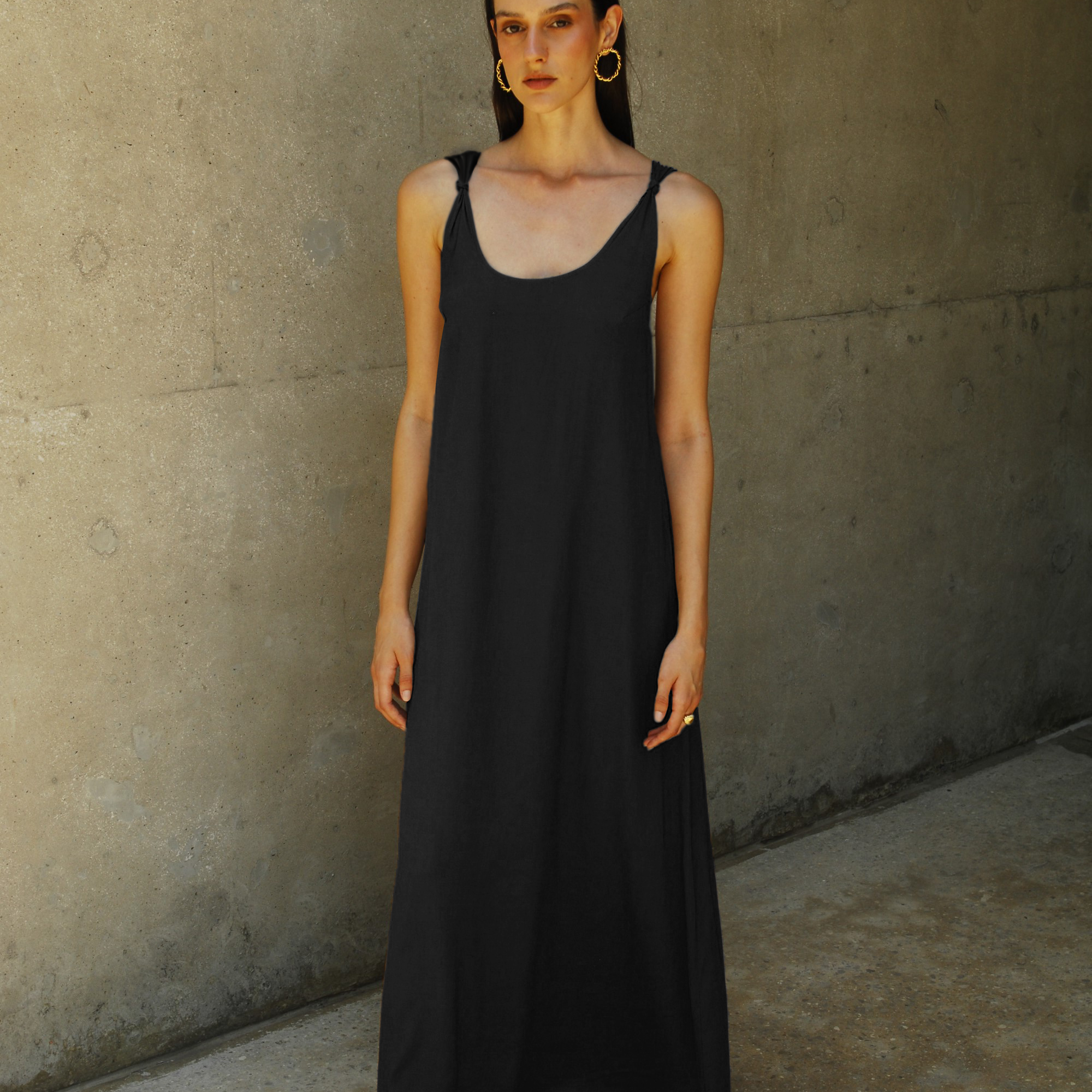 A model wearing a black maxi dress with a deep neckline and knot straps, standing against a concrete background.