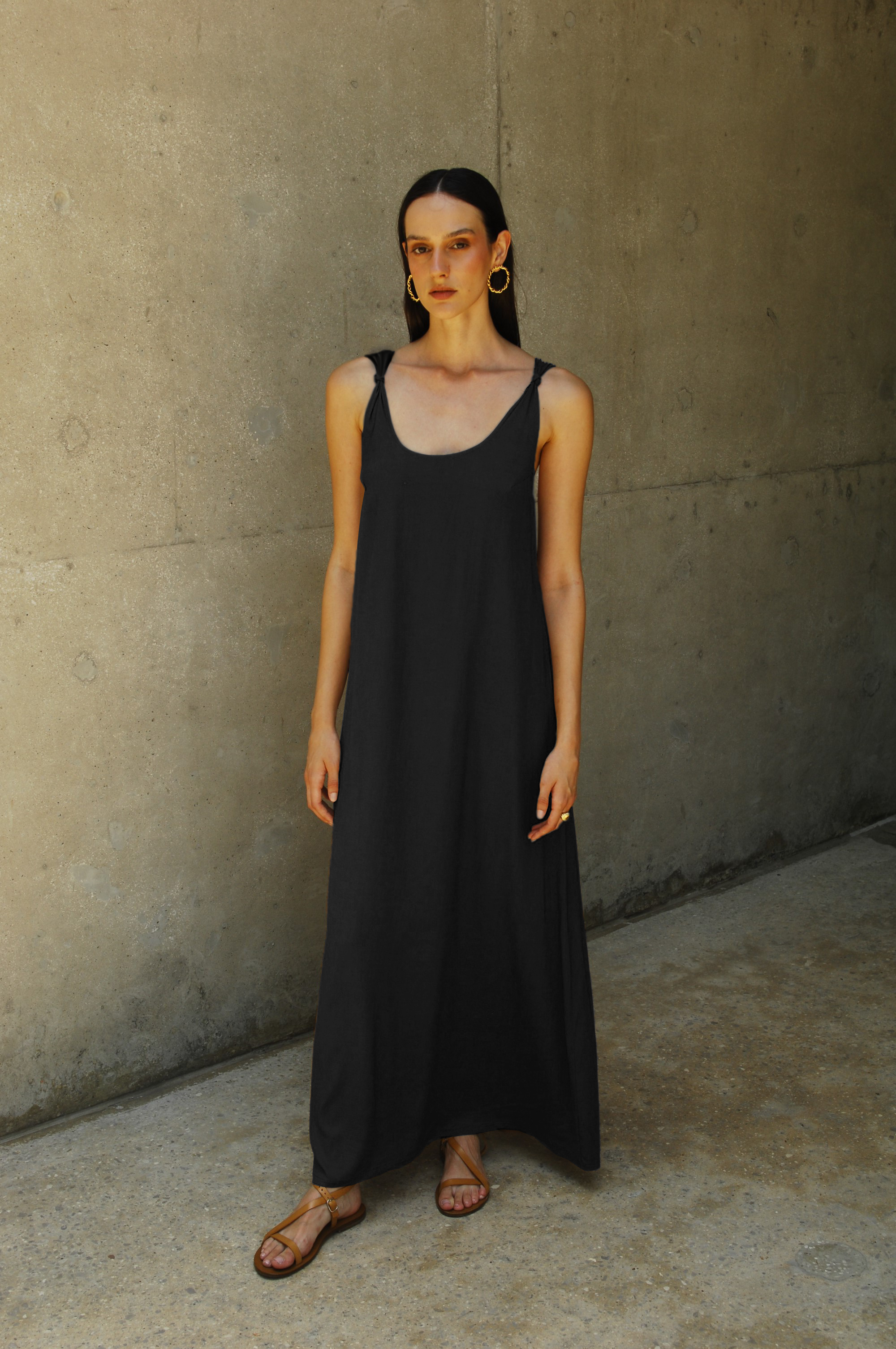 A model wearing a black maxi dress with a deep neckline and knot straps, standing against a concrete background.