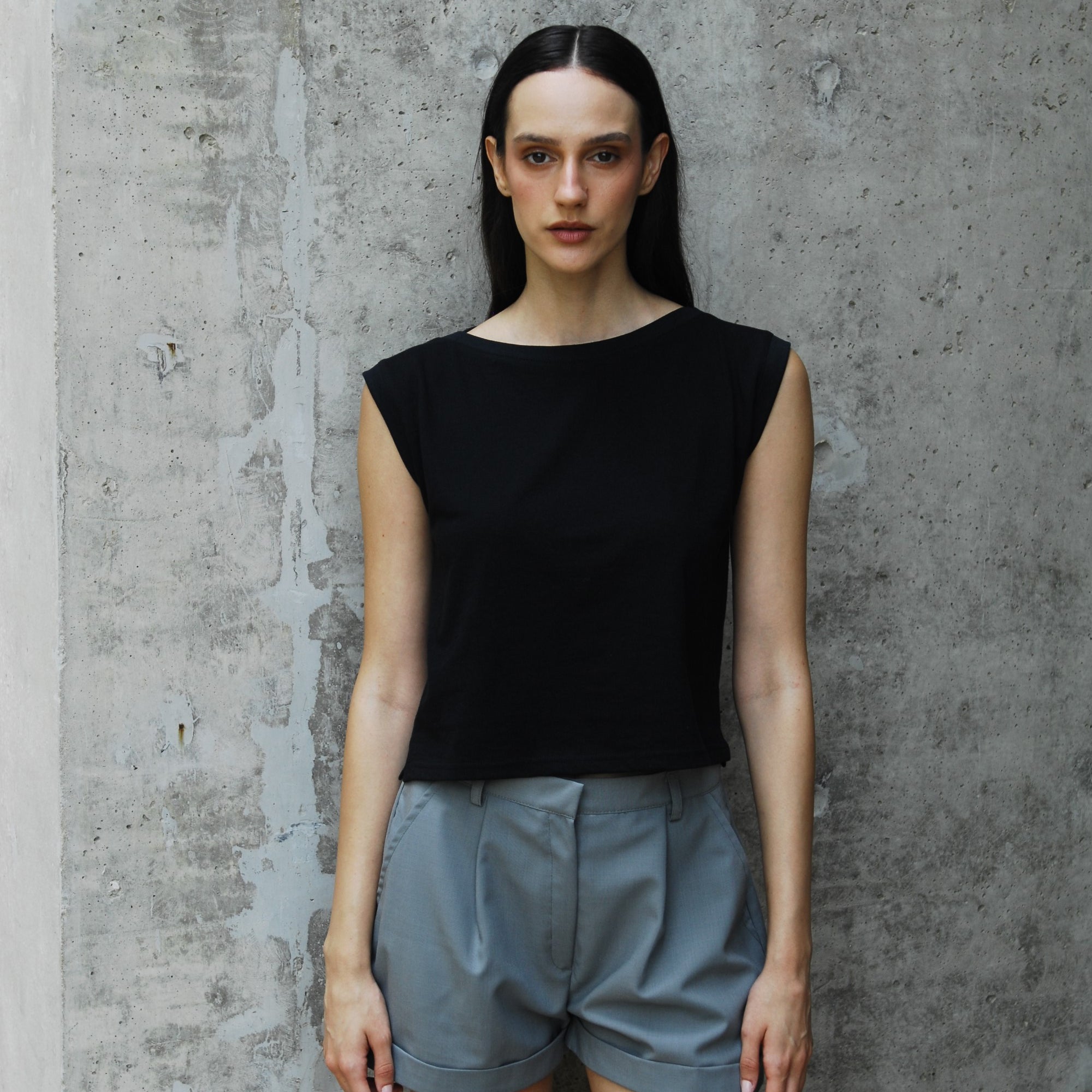 A woman standing against a concrete background wearing a black dahlia ishirt with a sailor's neckline and light gray shorts.