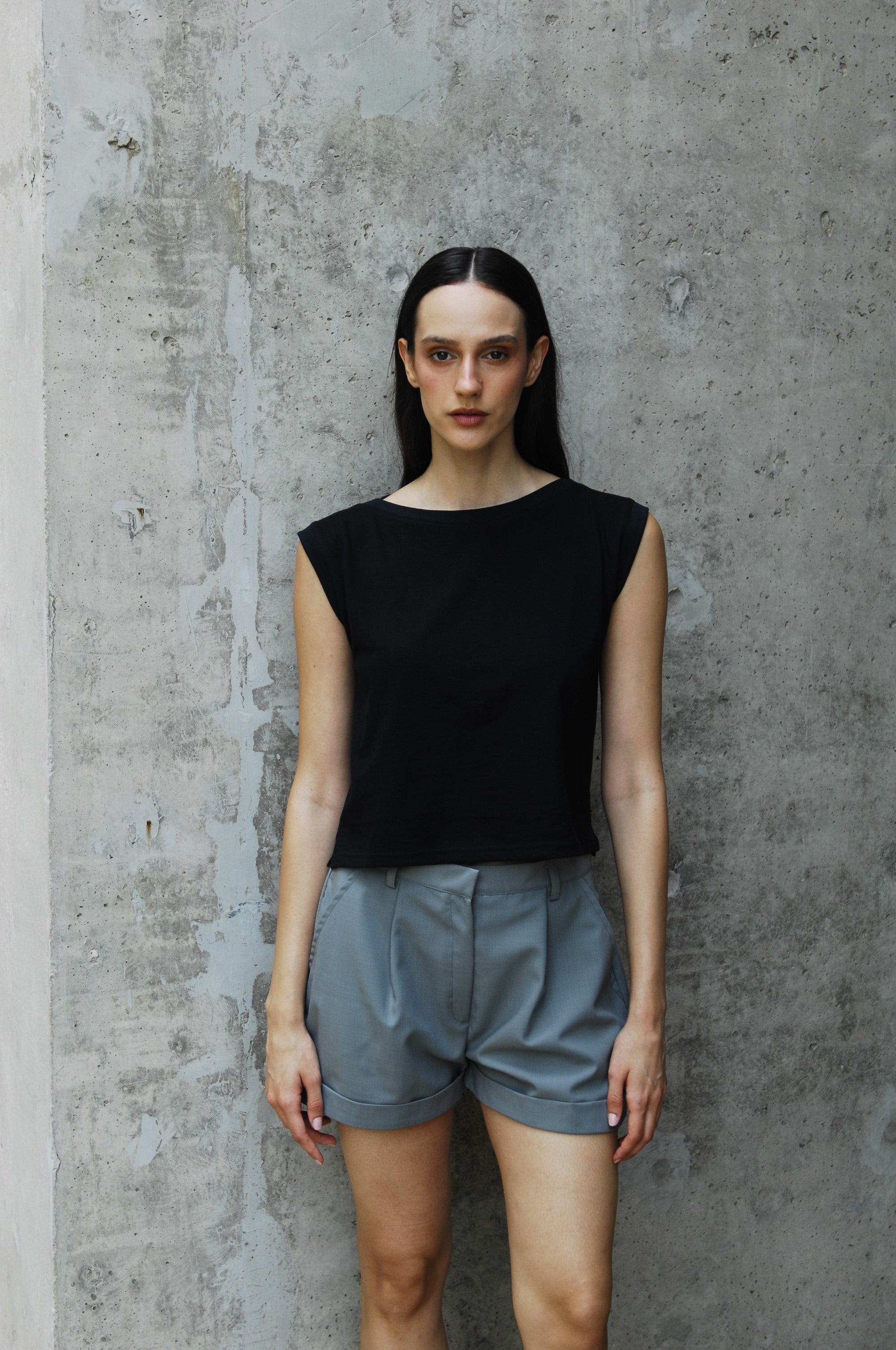 A woman standing against a concrete background wearing a black dahlia ishirt with a sailor's neckline and light gray shorts.