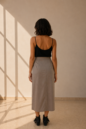 A woman stands with her back to the camera wearing a black spaghetti-strap camisole and a high-waisted, knee-length taupe-gray skirt, set against a warm beige wall with soft window shadow patterns.