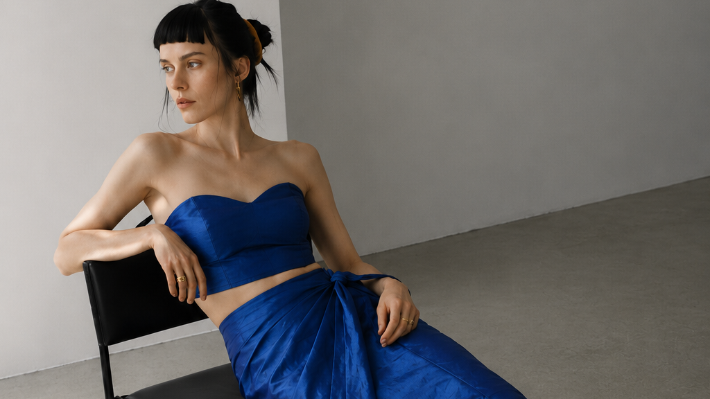 A woman models a vibrant royal blue two-piece set consisting of a strapless bandeau top and a matching wrap-style midi skirt, seated on a black chair in a minimalist studio setting.