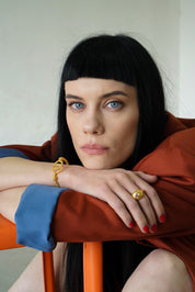 A woman with their elbow on a chair wearing a gold-plated farah ring with a blush pink stone on the finger.