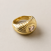 A gold ring with a ribbed, wing-like textured band and a central oval pale pink gemstone, shown on a neutral light beige background.
