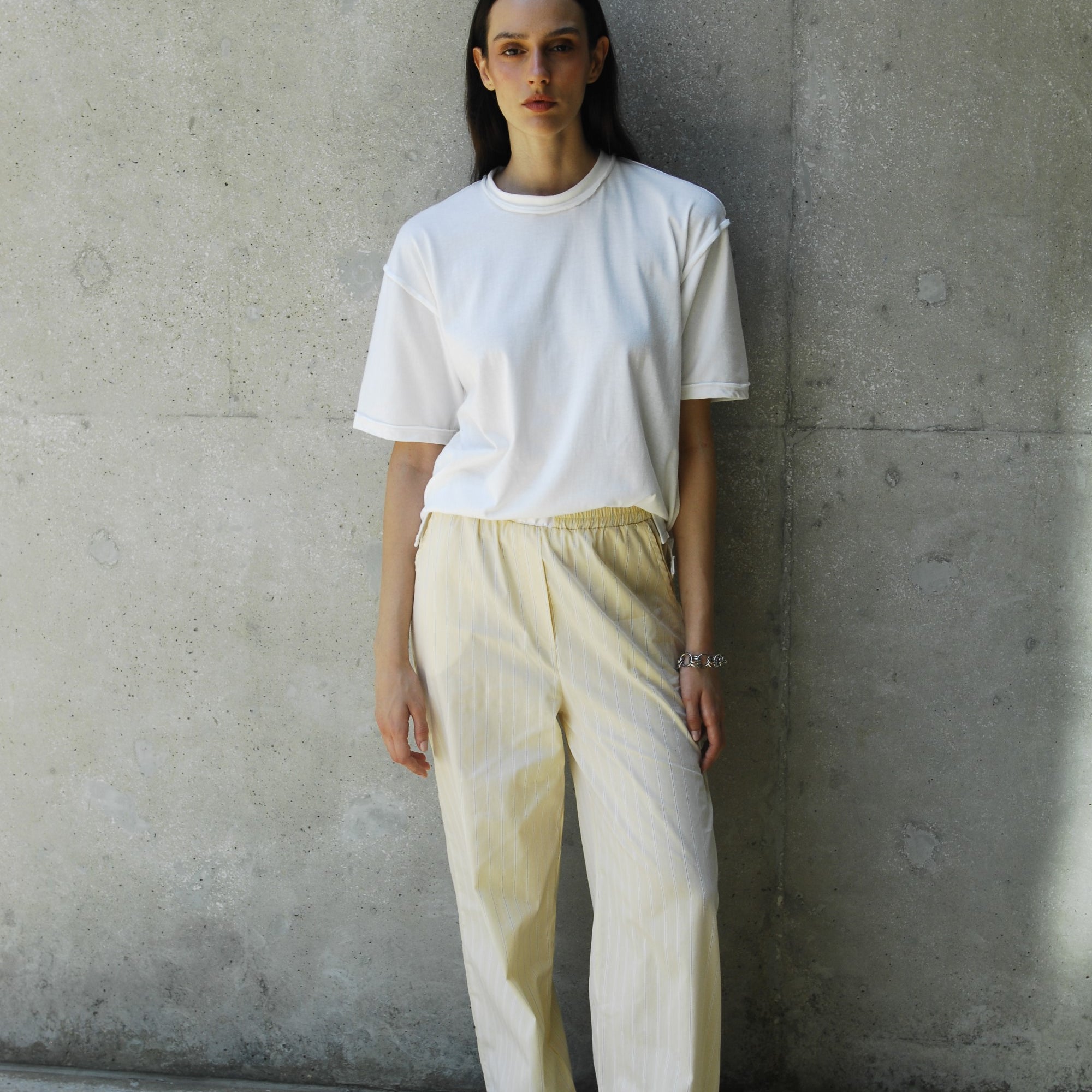 A woman wearing the sana white relaxed fit t-shirt and beige pants, standing against a concrete wall.
