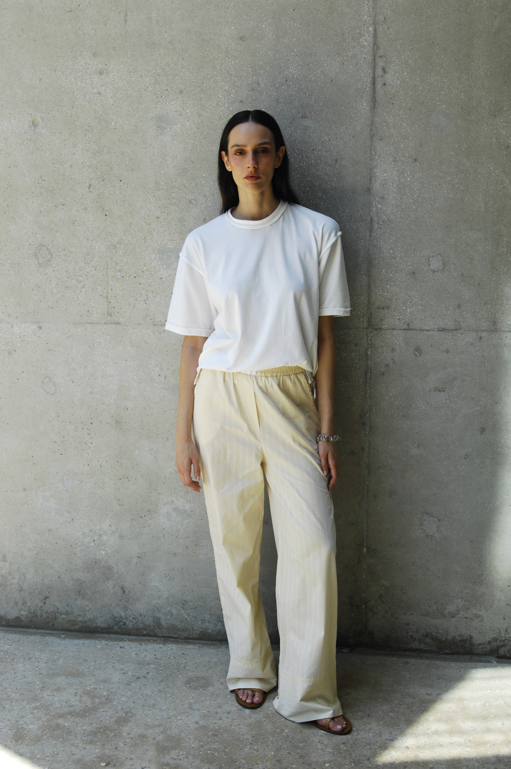 A woman wearing the sana white relaxed fit t-shirt and beige pants, standing against a concrete wall.