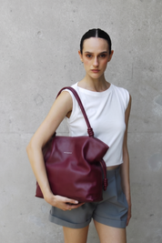 Woman holding Rumi burgundy handbag against a gray concrete wall