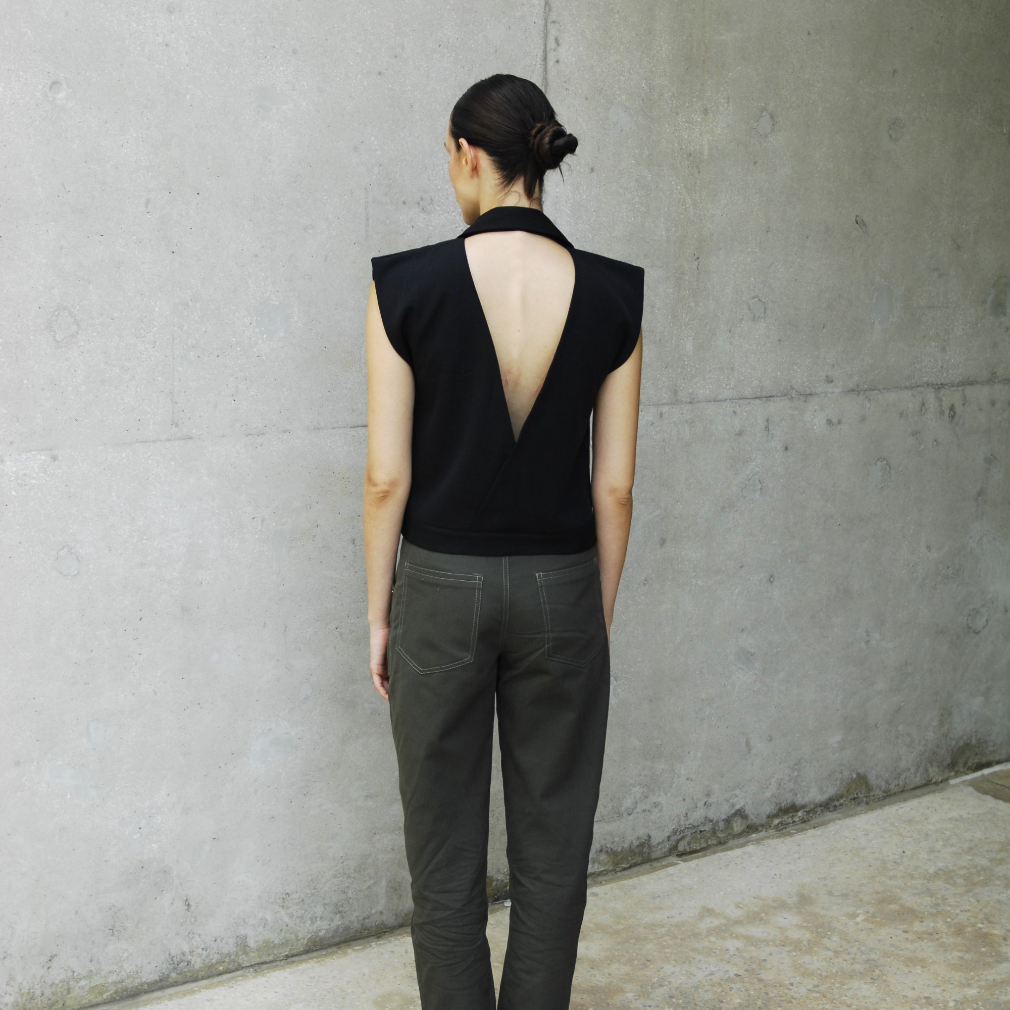 woman wearing a black backless vest and khaki pants standing on a concrete floor.