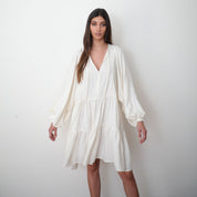 A woman standing against a white background, wearing a white midi dress with long puffy sleeves and a ruffled skirt.